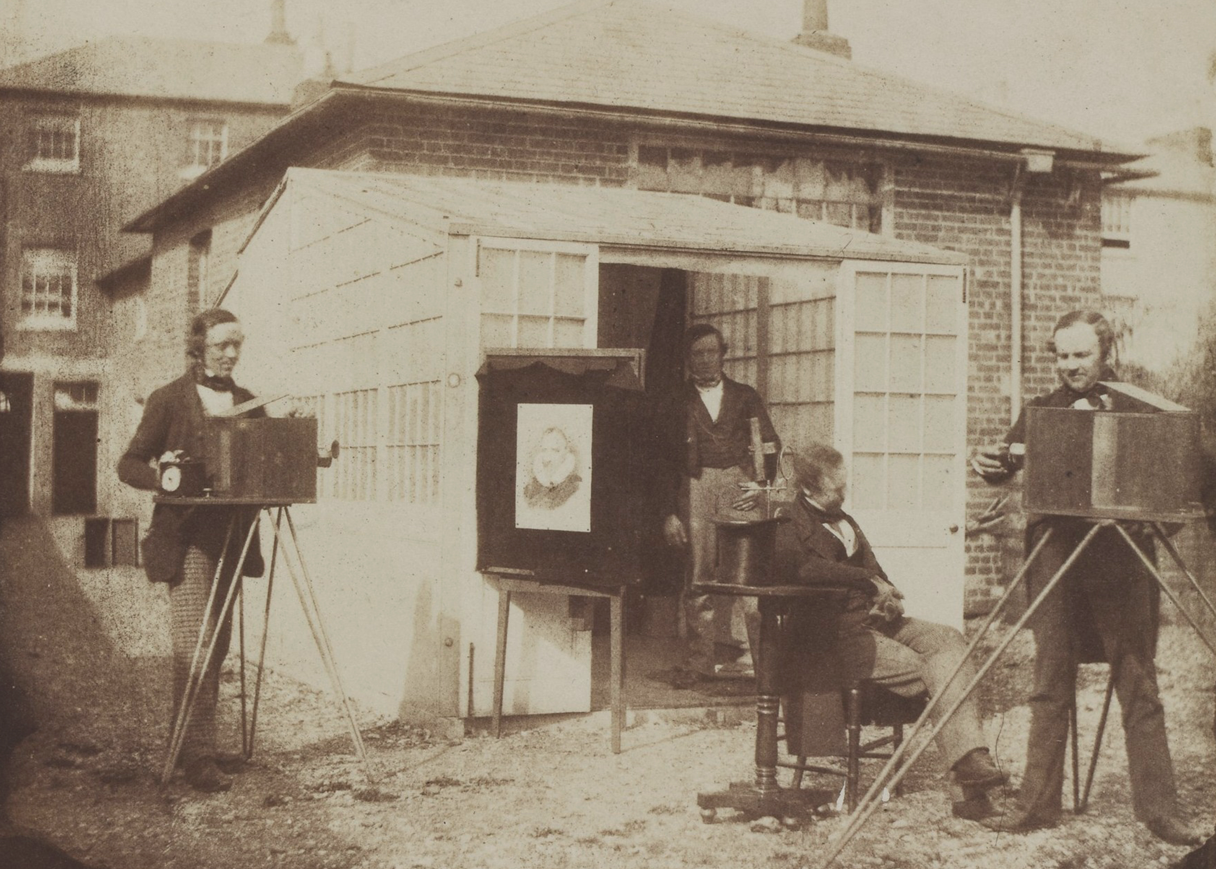 Four men stand outside a 19th-century photo studio. Two hold large box cameras on tripods, while another is seated. A portrait is displayed nearby.